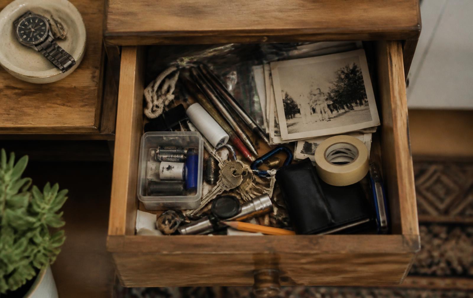 An open wooden drawer holding keys, pens, batteries and old photographs